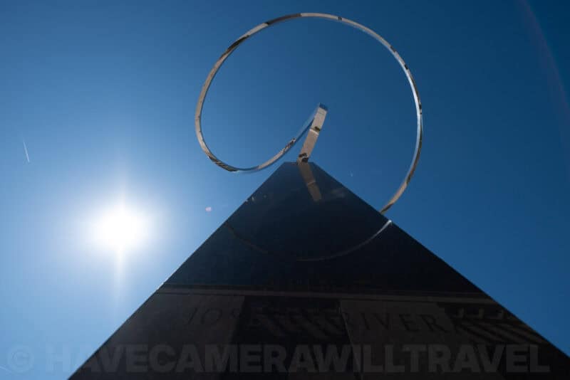 Spiral steel sculpture atop a dark pyramid monument against blue sky with sun flare