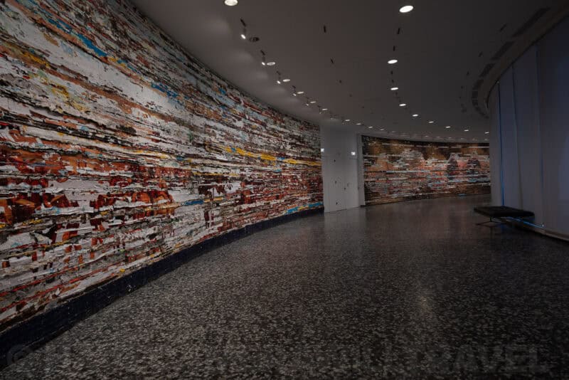 Curved museum corridor lined with a large abstract mural made of layered strips of color