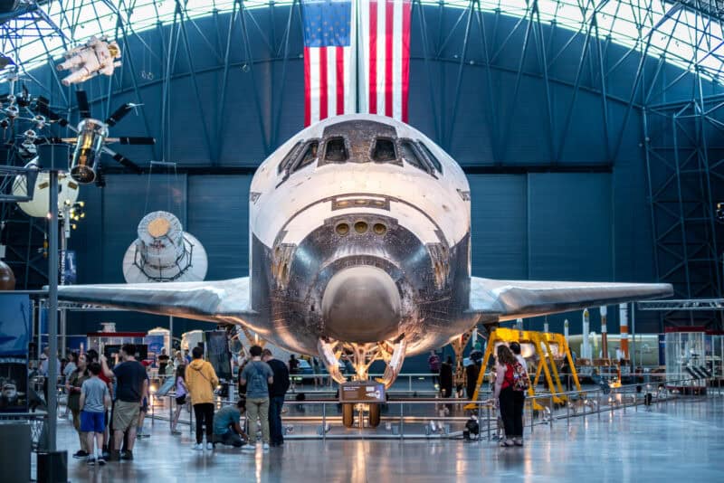 Space shuttle nose on display beneath a large American flag inside a museum hangar