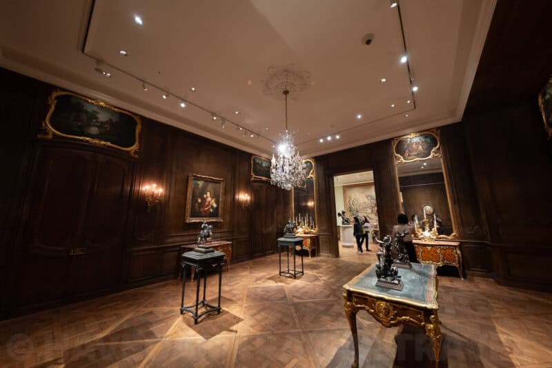 Ornate wood-paneled gallery with gilded furniture, chandelier, and framed paintings on the walls