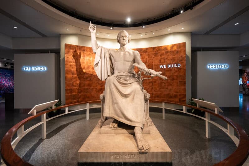Seated marble statue of George Washington pointing upward in a museum rotunda display