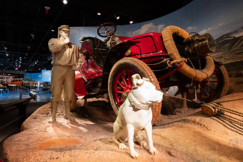 Museum diorama of a man, dog, and antique red automobile on a rocky terrain display