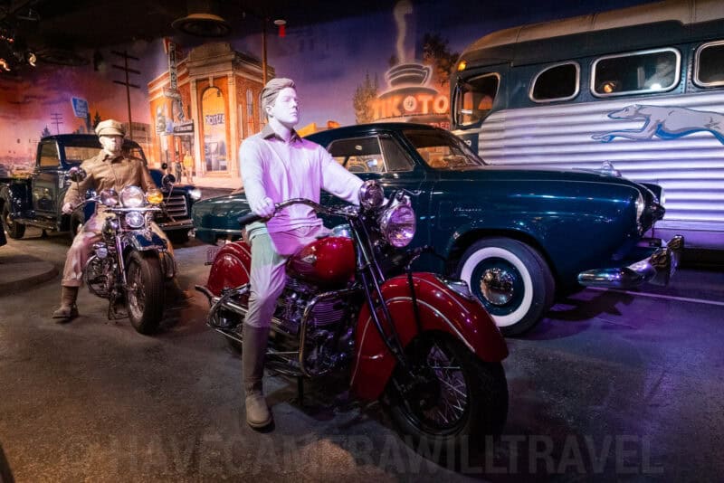 Mannequins on vintage motorcycles beside classic cars in a retro transportation museum display