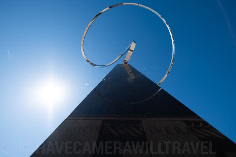 Spiral metal sculpture atop a dark pyramid monument silhouetted against bright sun