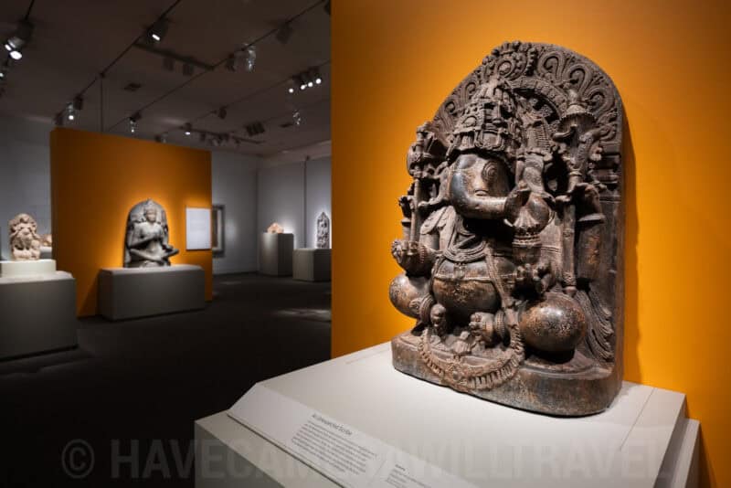 Carved stone sculpture displayed against an orange museum wall with other artifacts in background