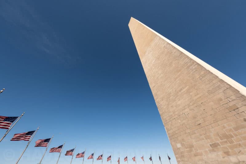 Washington Monument's sharp stone apex against blue sky with American flags below