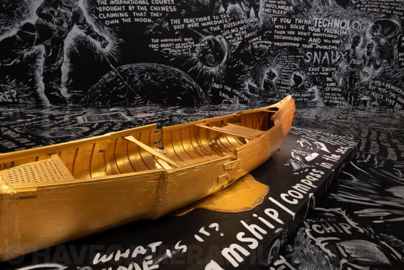 Gold-painted canoe displayed on a chalkboard-illustrated museum exhibit base