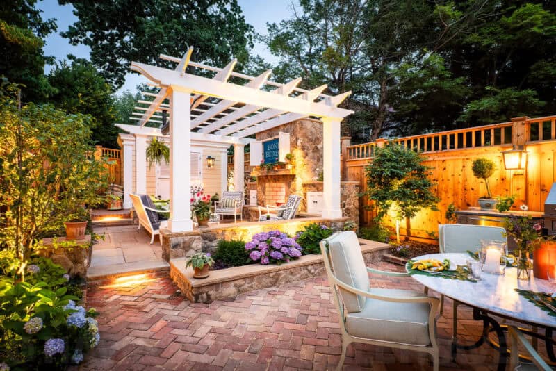 Backyard patio with white pergola, stone fireplace, and brick paving lit at dusk