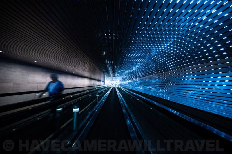 Blurred figure riding a moving walkway through a glowing blue LED tunnel