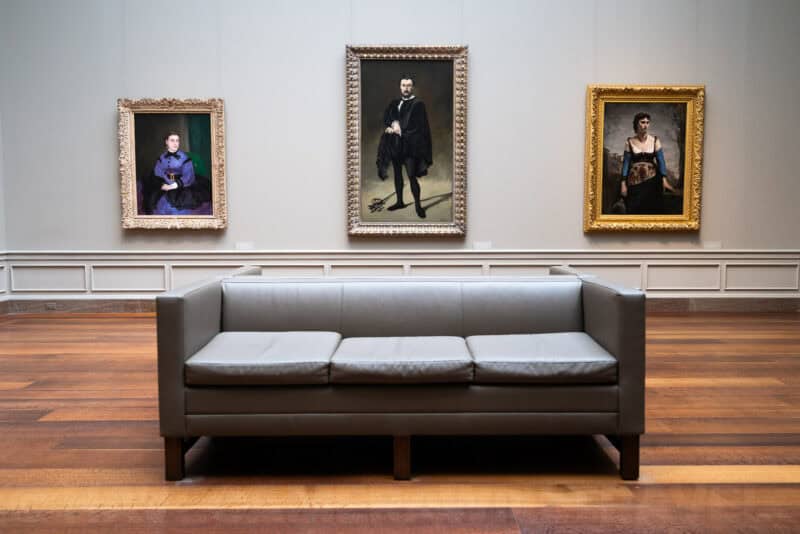 Gray leather sofa beneath three framed portrait paintings in a museum gallery