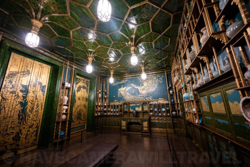 Ornate Peacock Room with green and gold walls, honeycomb ceiling lights, and shelved porcelain vases