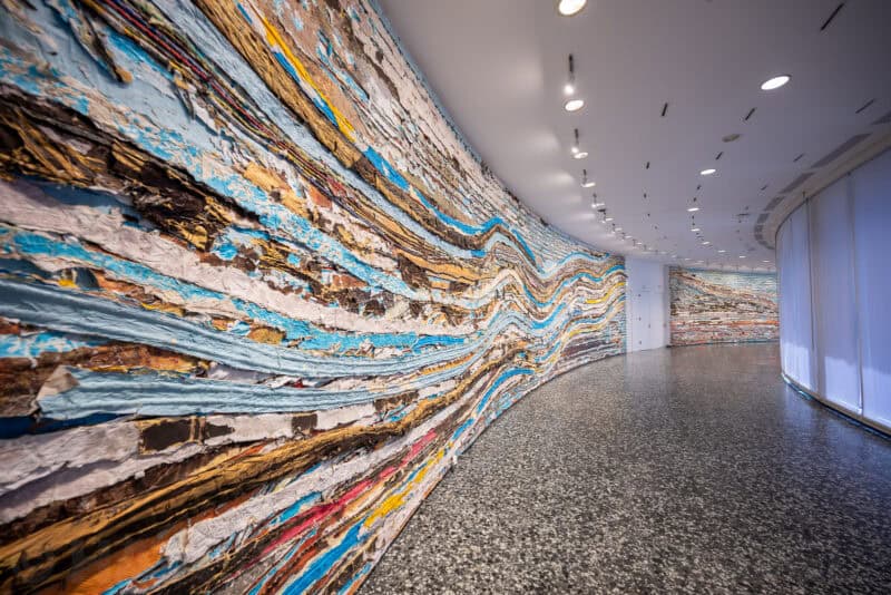 Curved museum gallery wall covered in a textured multicolored layered art installation