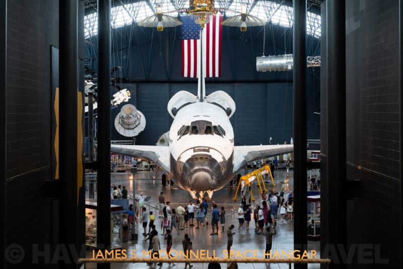 Space shuttle displayed nose-on beneath a hanging American flag in a museum hangar