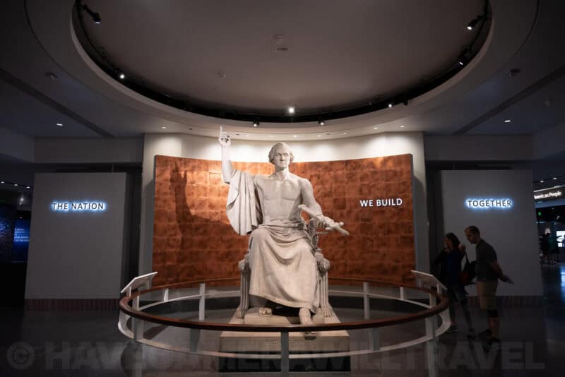 Seated marble statue of George Washington in a museum rotunda with a copper backdrop reading 'We Build'