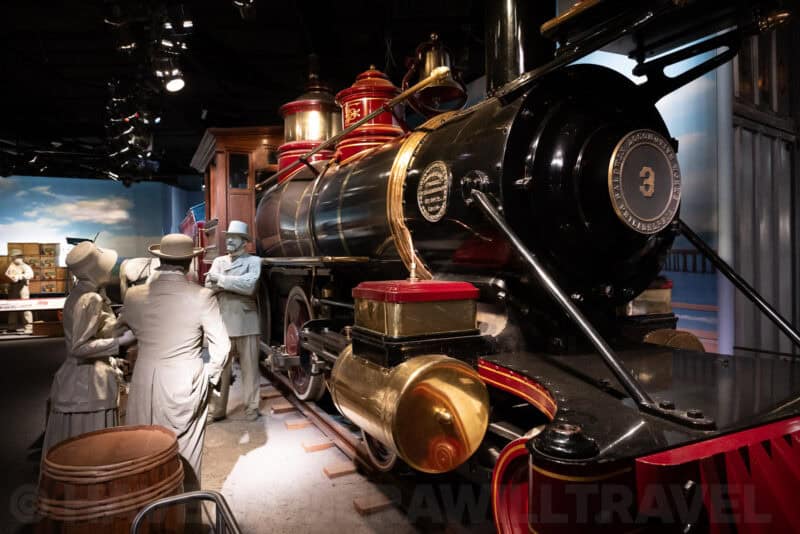 Vintage black steam locomotive with red trim on museum tracks beside period-costumed mannequins