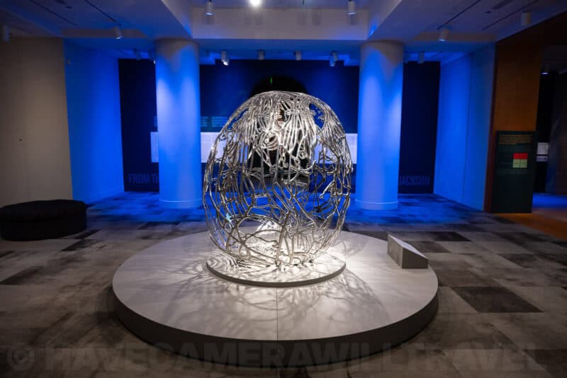 Egg-shaped wire sculpture lit on a circular platform in a blue-lit museum hall