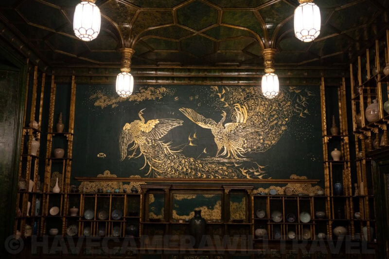 Gilded peacock mural on a dark wall inside an ornate room lined with shelved porcelain