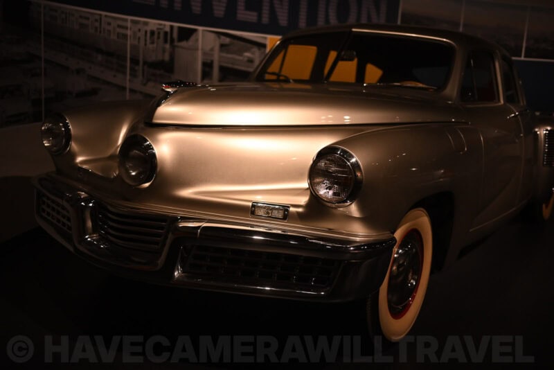 Silver vintage Tucker automobile with chrome grille and whitewall tire under a dim museum spotlight