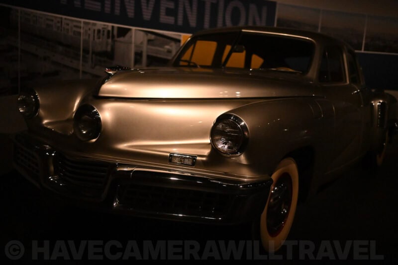 Classic silver Tucker car front end photographed in near-darkness beneath a museum spotlight