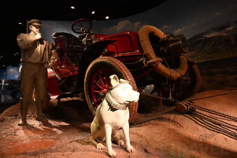 Museum diorama of a vintage red car, driver figure, and white dog statue in a dim high-ISO test shot