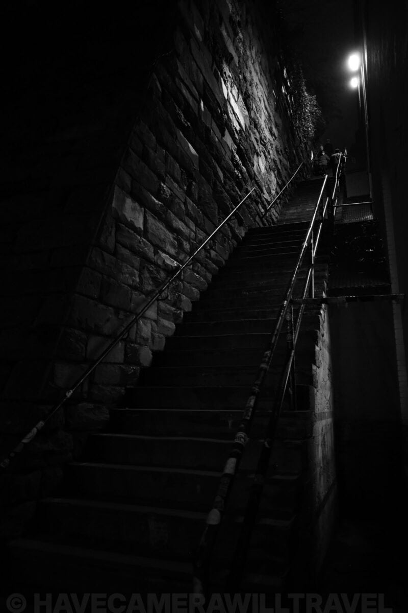 Stone staircase along a rough-cut rock wall lit by a single lamp at the top at night