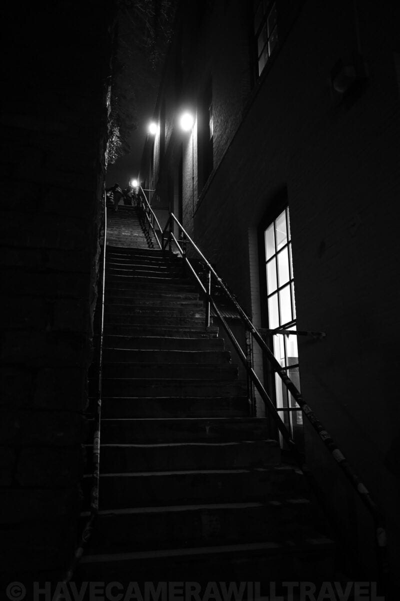 Dimly lit outdoor staircase between brick buildings climbing toward distant streetlights at night