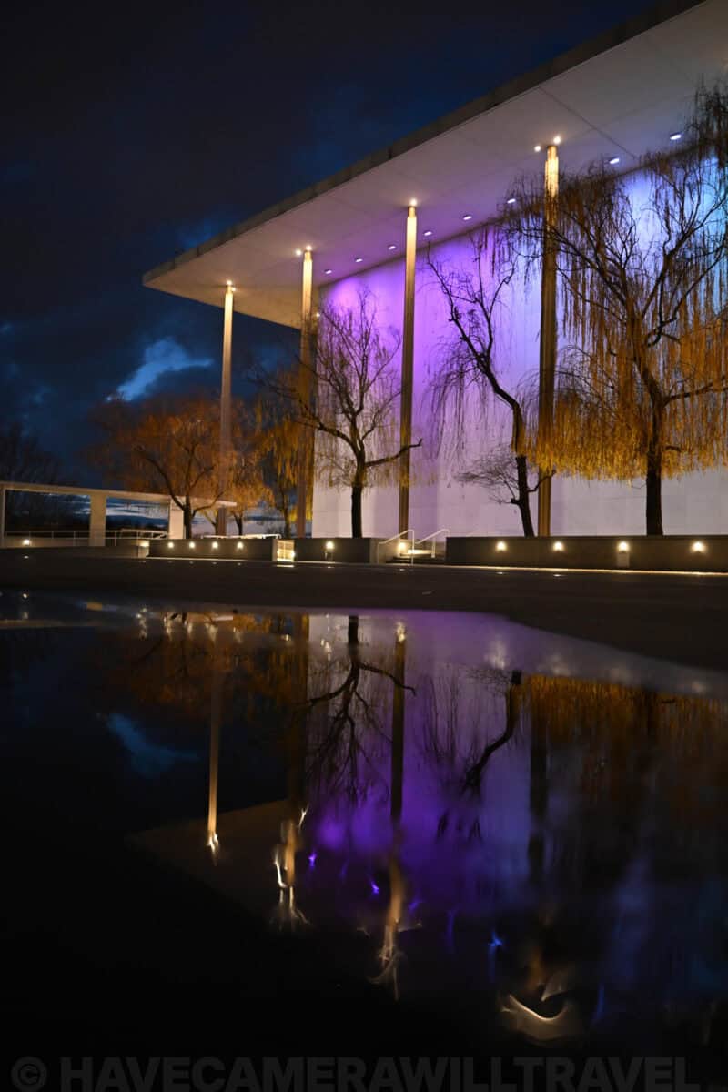 Kennedy Center terrace with purple-lit colonnade and golden willow trees reflected in a still pool at night