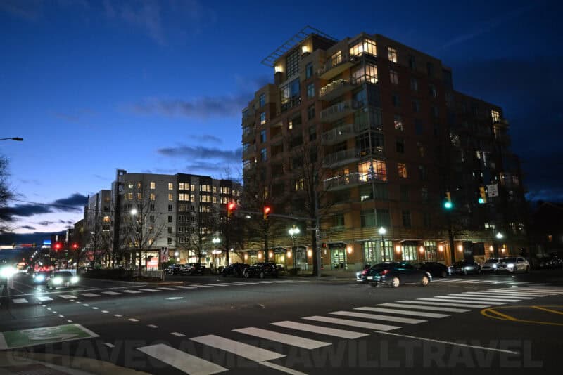 Illuminated apartment building on a city street corner at dusk with cars at an intersection