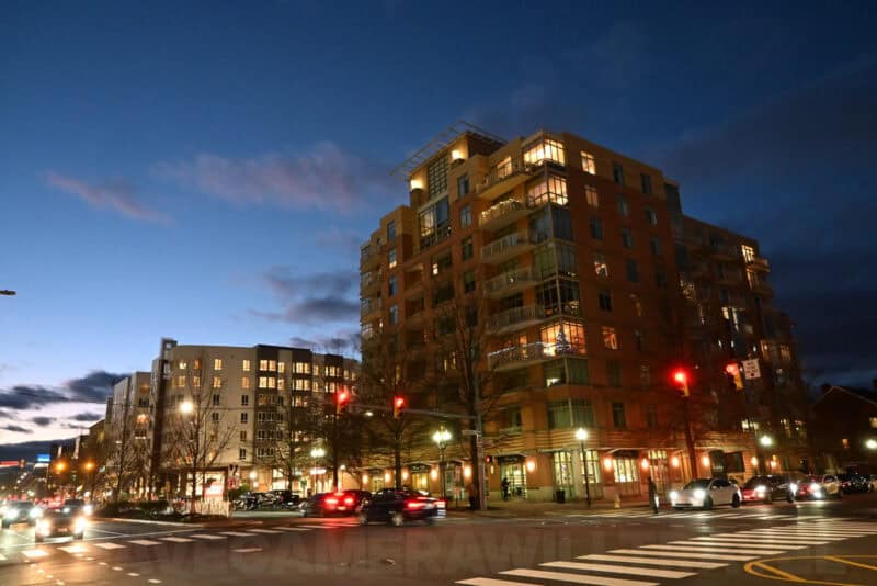 City street corner at dusk with a lit apartment building and traffic waiting at a red light