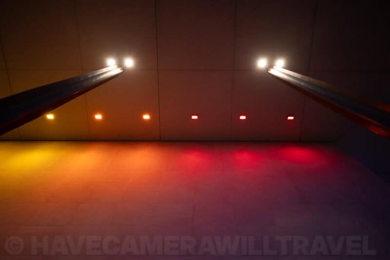Colored floor lights in orange and red glowing beneath angled overhead beams in a dark hall