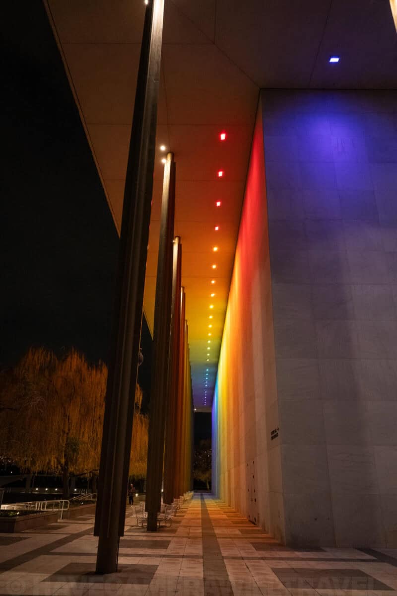 Covered walkway with rainbow-lit wall and colored light installation at night