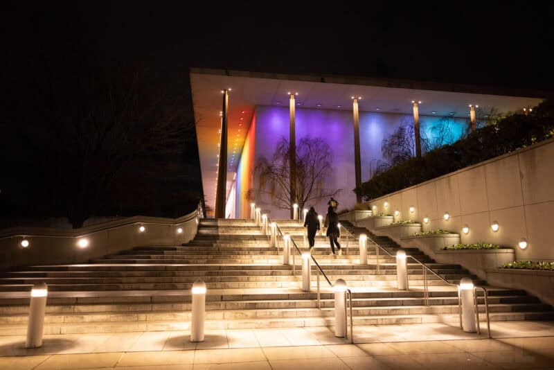 The Kennedy Center's illuminated staircase and rainbow-lit terrace at night with visitors descending