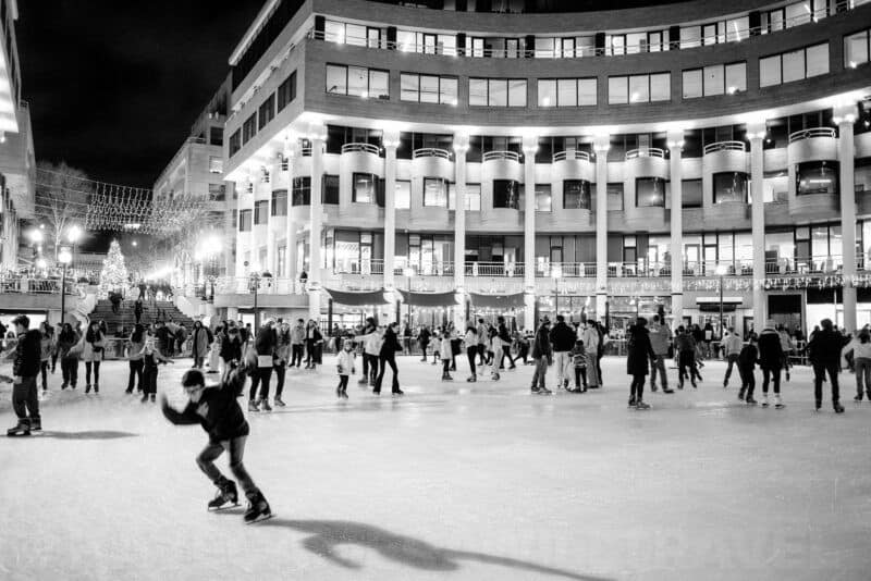 Black and white night shot of an outdoor ice rink with skaters in front of a colonnaded building