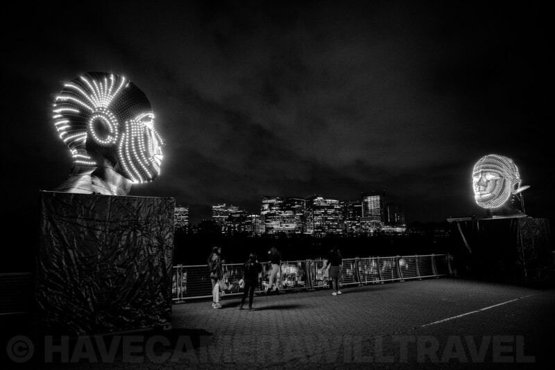 Illuminated LED sculpture heads facing a lit city skyline at night in black and white