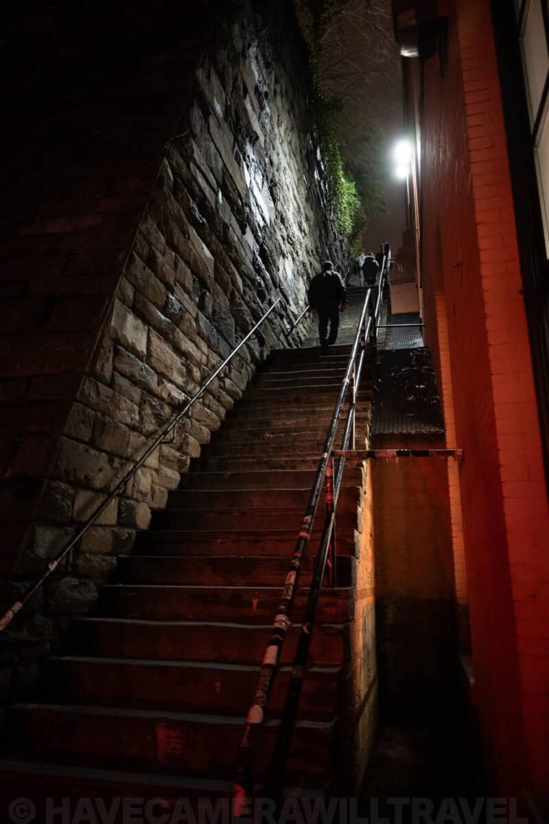 A person climbing a steep stone staircase between illuminated walls at night