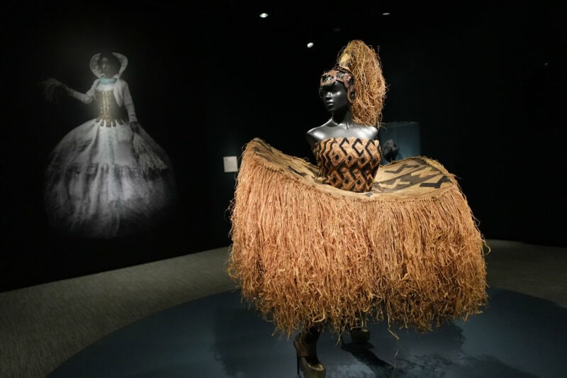 Mannequin in a raffia grass skirt and headdress displayed before a ghostly gown backdrop in a dark museum gallery
