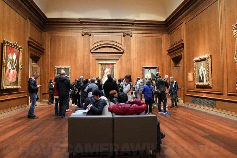 Museum visitors gathered around framed portrait paintings in a wood-paneled gallery room