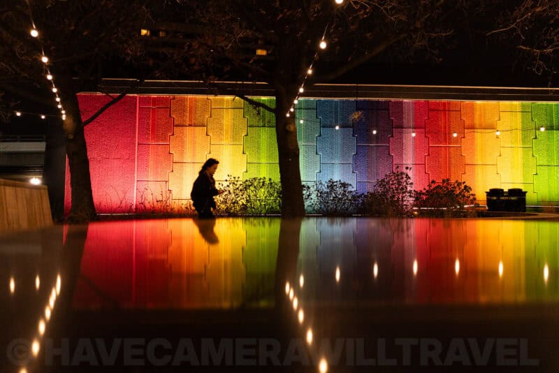 Silhouetted figure walking past a rainbow-lit wall reflected in water at night