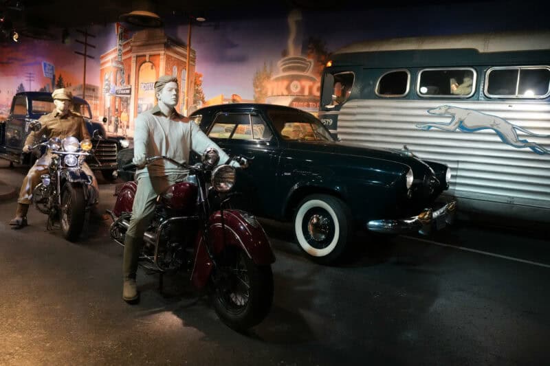 Vintage roadside diorama with mannequins on motorcycles beside a classic car and Greyhound bus
