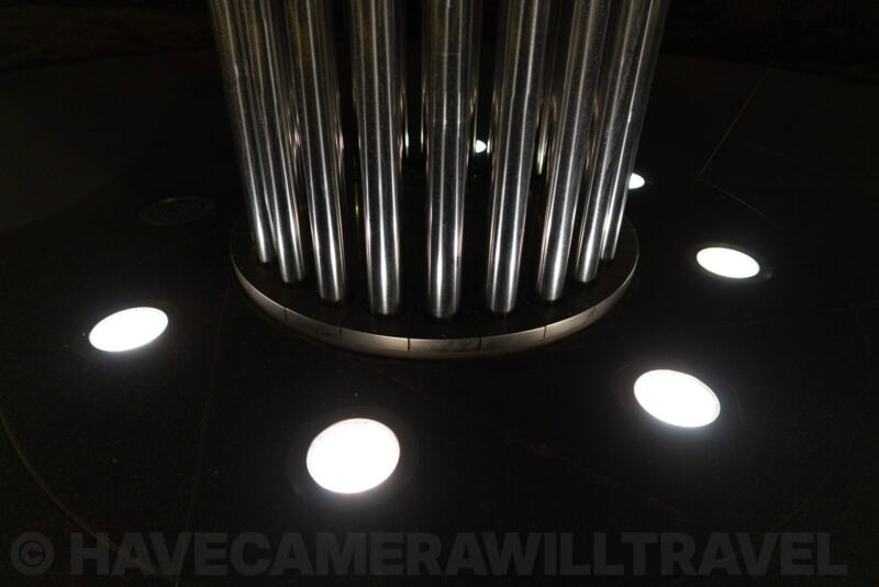 Close-up of a tall metal sculpture's base ringed by round floor lights at night