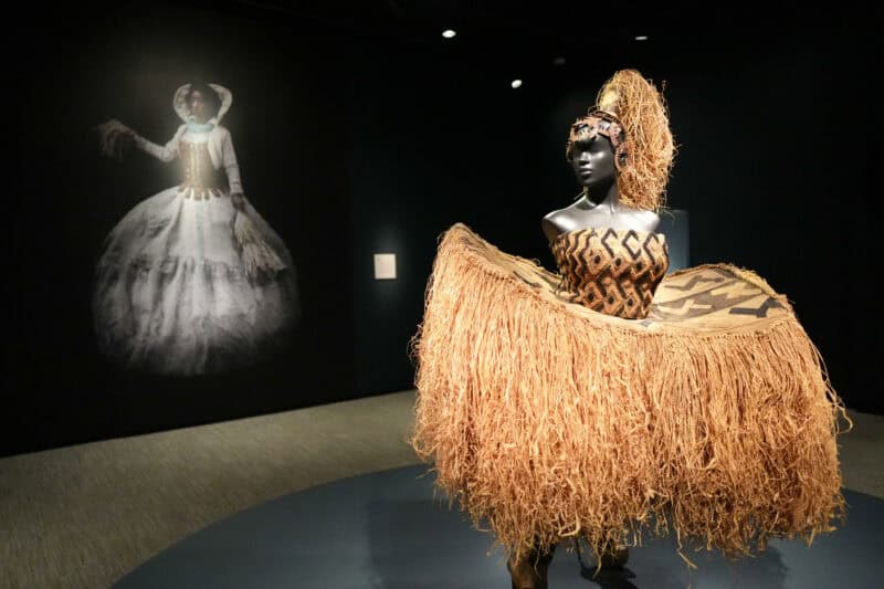 Museum figure in a raffia grass skirt costume standing before a projected historical gown image