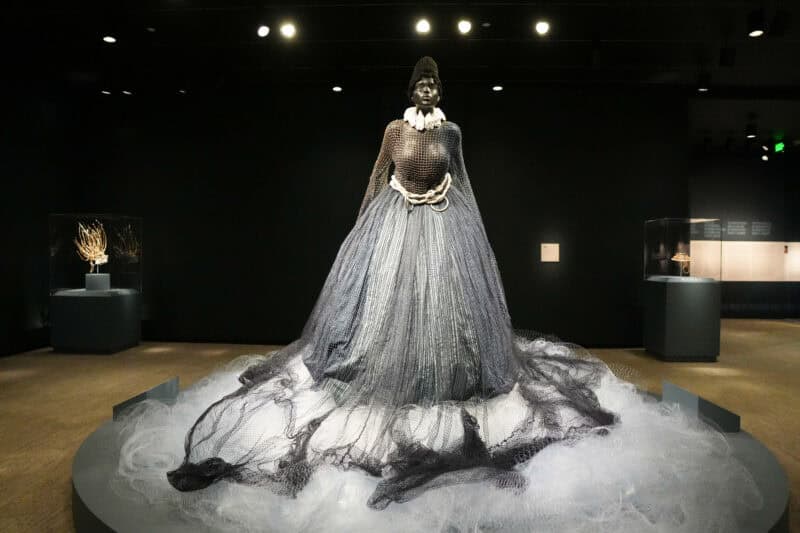 Figure in a flowing metallic netted gown displayed on a round platform in a dim gallery