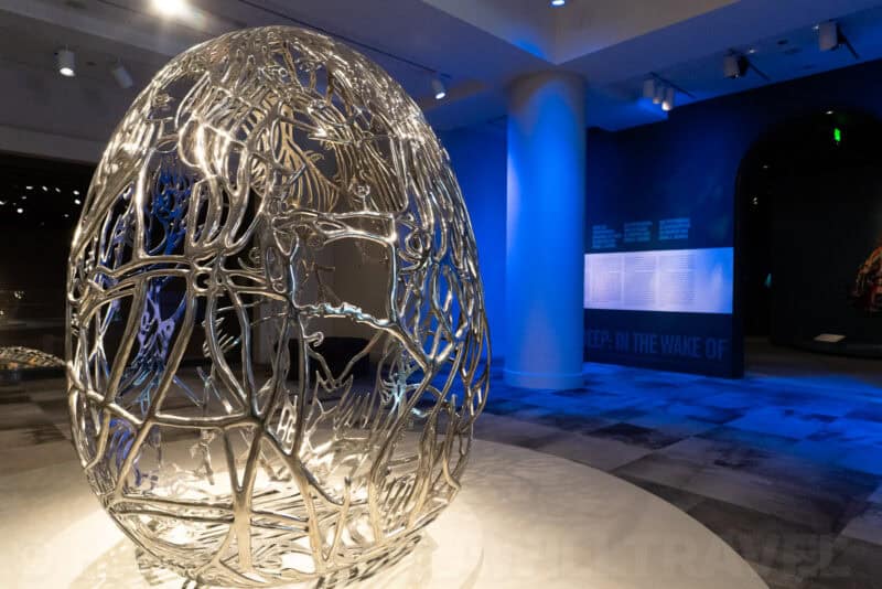 Large silver wire-sculpted egg artwork lit blue in a museum exhibit hall