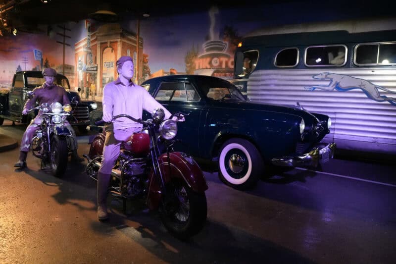 Purple-lit vintage scene with mannequins on motorcycles beside a classic car and bus mural