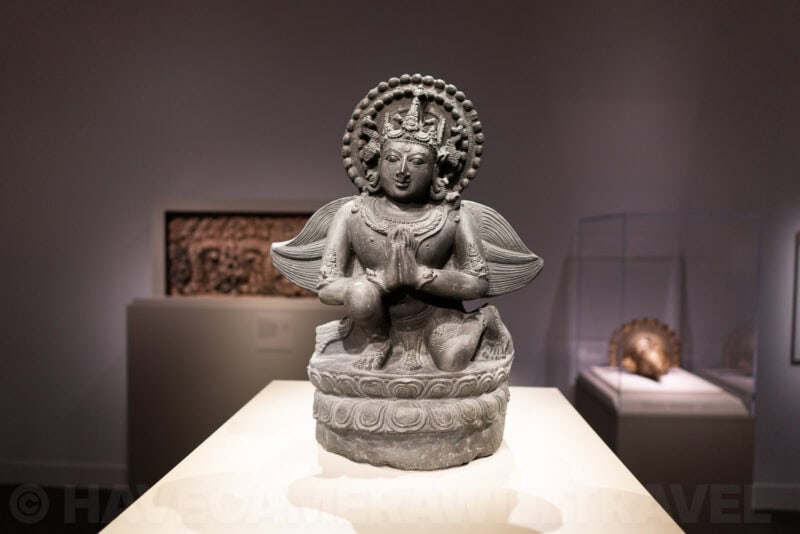 Small stone statue of a kneeling deity with wings displayed on a museum pedestal