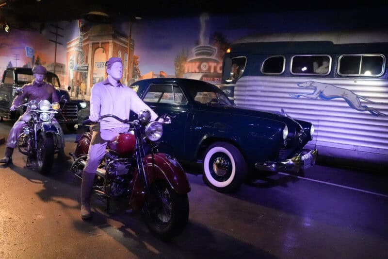 Purple-lit vintage roadside diorama featuring mannequins on motorcycles and a classic car