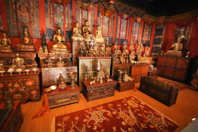 Tibetan Buddhist shrine room filled with bronze statues, painted cabinets, and a patterned rug