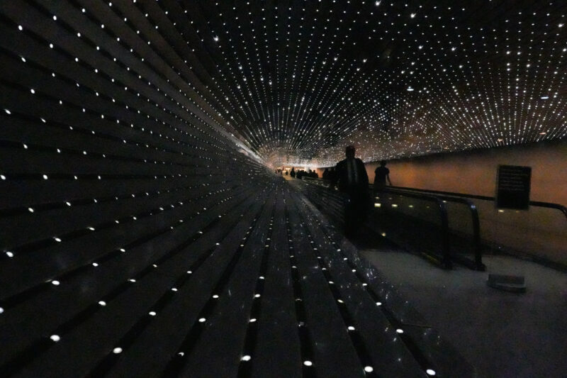 Tunnel-like moving walkway covered in twinkling lights with silhouetted passengers riding through