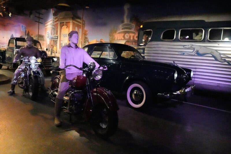 Purple-lit vintage diorama with mannequins on motorcycles beside a classic car and bus mural