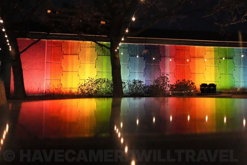 Rainbow-lit wall reflected in a pool of water at night beside silhouetted trees